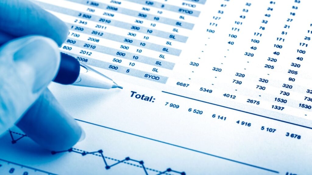 Close-up of a hand holding a ballpoint pen reviewing financial statements with rows of numerical data and totals, symbolizing financial reporting, accounting accuracy, and data-driven decision-making.